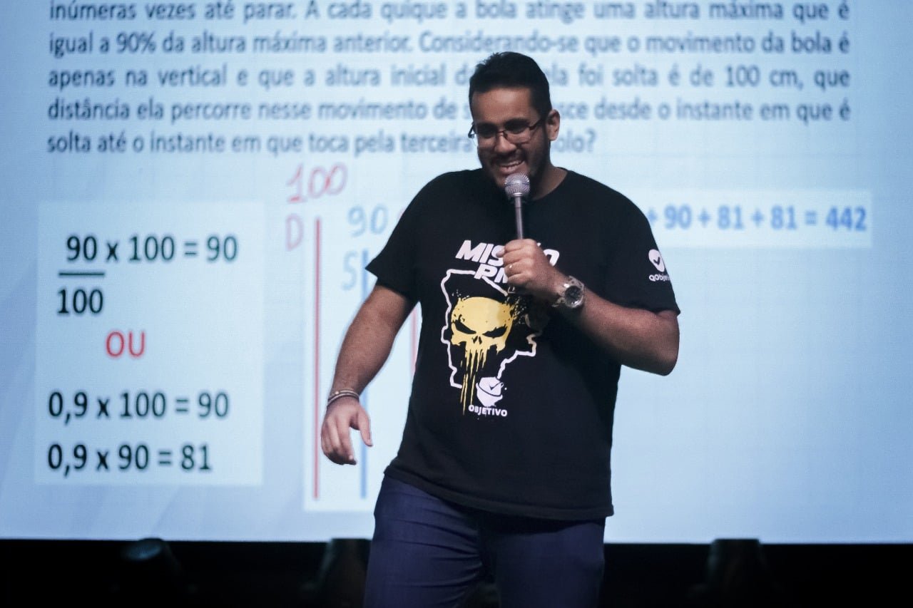 Rafael Cardoso dando aula de matemática num palco, resolvendo questão no telão